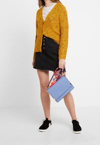 Cardigan lavorato a maglia giallo senape, gonna di jeans nera con bottone frontale, borsa blu con tracolla decorata, sneakers nere.