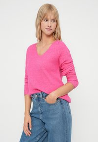 Różowy sweter w serek z długimi rękawami, z teksturowanym splotem. W połączeniu z niebieskimi dżinsami z wysokim stanem, co tworzy casualowy wygląd.
