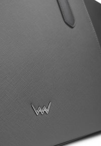 Graue Handtasche mit Textur, die einen glatten Ledergriff und ein silbernes Logo-Detail auf der Vorderseite aufweist.