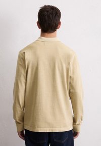 Beżowa, długorękawowa koszulka polo z kołnierzykiem, o prostym wzorze i gładkiej fakturze, prezentowana z tyłu na jasnym tle.