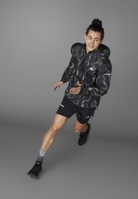 Schwarze und graue Tarnjacke mit Kapuze, kombiniert mit schwarzen Shorts und Laufschuhen. Die Socken sind mit Streifen und Markenlogos versehen.