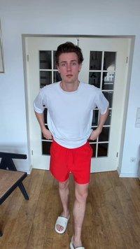 Jeune homme en t-shirt blanc, short rouge et chaussons debout à l'intérieur sur un sol en bois devant des portes doubles vitrées.