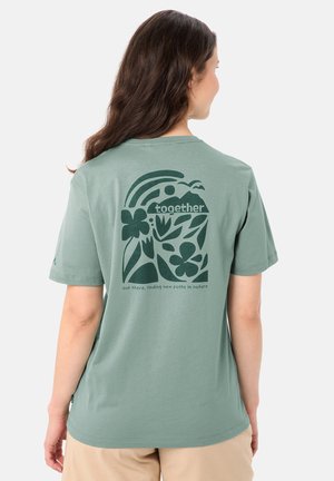 Vrouw met lang bruin haar, gekleed in een lichtgroen T-shirt met een donkergroene natuurthema-afbeelding en het woord "samen" op de achterkant.
