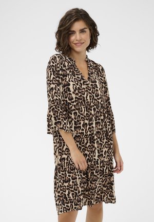 Femme aux cheveux courts et bouclés portant une robe léopard mi-longue avec des manches cloche trois-quarts, debout devant un fond blanc uni.