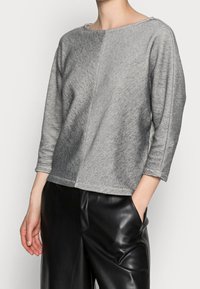 Harmaa raidallinen paita veneen kaula-aukolla, kolmanneksen pituisilla hihoilla ja sivurei'illä, yhdistetty mustiin tekonahkahousuihin. Sileä tekstuuri ja moderni muotoilu.