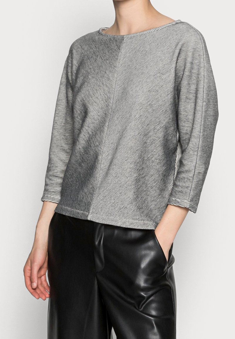 Harmaa raidallinen paita veneen kaula-aukolla, kolmanneksen pituisilla hihoilla ja sivurei'illä, yhdistetty mustiin tekonahkahousuihin. Sileä tekstuuri ja moderni muotoilu.