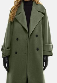 Oliven grøn, dobbeltradet frakke med bred krave, store knapper og opretstående manchetter. Båret over en sort turtleneck og læderhandsker.