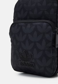 Gros plan sur la poche avant d'un sac à dos noir Adidas avec un motif de logo trèfle ton sur ton et une tirette de fermeture éclair.