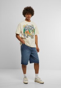 Beige grafisk t-skjorte med et campingmotiv som viser trær og dyr, kombinert med blå denimshorts, hvite sokker og beige joggesko.