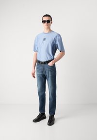 Light blue short-sleeve shirt with dark blue accents, paired with blue jeans and black lace-up shoes. Model wears sunglasses.
