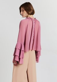 Donna in blusa rosa con bottoni sul retro e maniche a campana sovrapposte, abbinata a pantaloni larghi beige, posata di fronte a uno sfondo semplice.