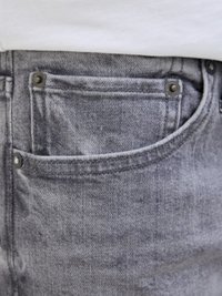Grå denim jeans med en frontlomme med synlige sømme og metalnitter, vist mod en lyse farvet stofbaggrund.