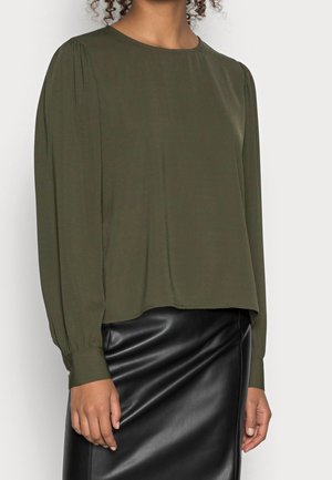 Mujer con una blusa de manga larga de color verde oliva con hombros abullonados y una falda de cuero negra.