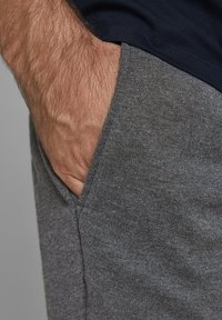 Šedé bavlněné tepláky s bočním kapsou, textura vypadá měkce a látka má jemný melírovaný vzor. Žádné viditelné logo.