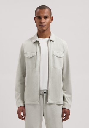 Man draagt een lichtgrijs jack met rits en borstzakken over een wit shirt en bijpassende lichtgrijze broek, staand tegen een effen achtergrond.
