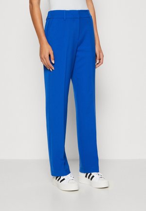 Persona que lleva pantalones azul brillante de corte recto y zapatillas blancas con rayas negras sobre un fondo liso.