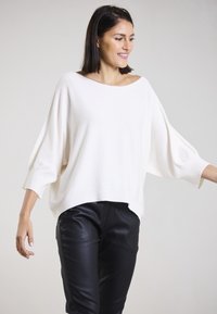 Weißer, oversized Pullover mit einem weiten Bootsausschnitt und Dreiviertelärmeln, kombiniert mit schwarzen, schmalen Hosen. Weiches Strickmaterial.