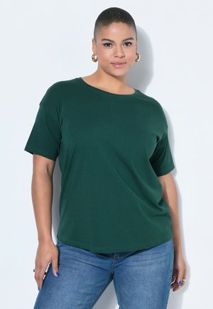 Frau mit kurzen Haaren, die ein grünes T-Shirt und blaue Jeans trägt, steht vor einem weißen Hintergrund mit entspannt an den Seiten hängenden Händen.