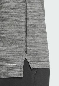 Sivi sportski top s teksturiranim završetkom, s bočnim prorezom i reflektirajućim logotipom. Materijal izgleda lagano i prozračno.