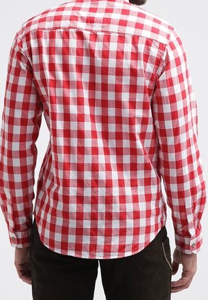 Homme portant une chemise à carreaux rouge et blanc à manches longues, vu de dos, avec un pantalon foncé sur un fond uni.