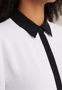 White shirt with a black, textured collar and placket, featuring a button at the collar, highlighting a contrast in color and material.