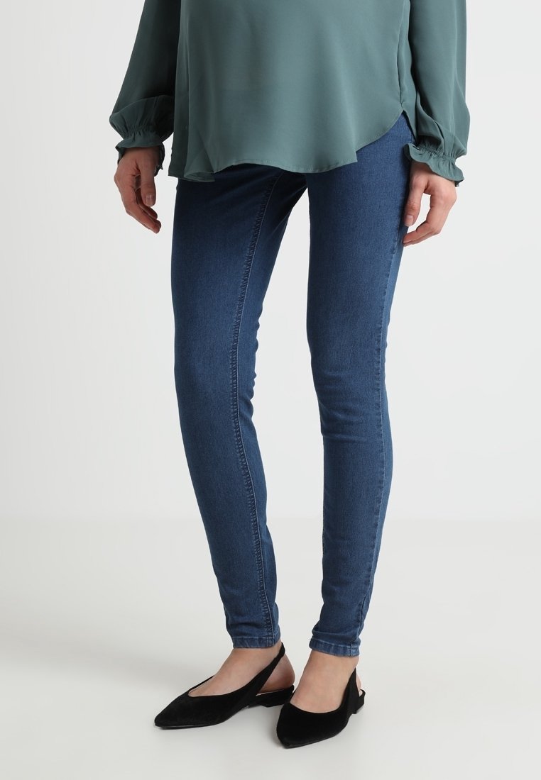 Jeans skinny in denim blu scuro, con una finitura liscia e una silhouette aderente, abbinati a scarpe nere con tacco basso e una blusa verde.