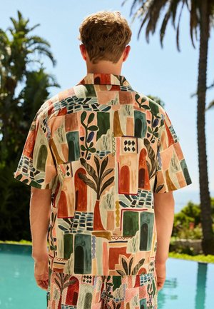 Mann steht am Pool, trägt ein kurzärmeliges Hemd und Shorts mit abstrakten geometrischen und Pflanzenmustern in Erdtönen.