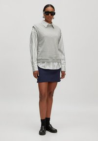 Grauer ärmelloser Sweatshirt über einem weiß gestreiften Hemd, kombiniert mit einem marineblauen Minirock, schwarzen Slippern und dunklen Socken.