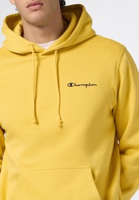 Felpa in cotone gialla con tasca frontale, cordini regolabili e logo "Champion" ricamato in blu sul petto. Tessuto morbido.