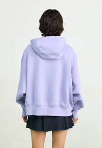 Moletom oversized lavanda com capuz ajustável, punhos e barra canelados. Usado sobre uma saia preta curta, destacando o ajuste relaxado e a textura.