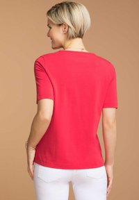T-shirt rossa a maniche corte realizzata in tessuto liscio, con scollatura arrotondata e vestibilità comoda, abbinata a pantaloni bianchi in un contesto neutro.