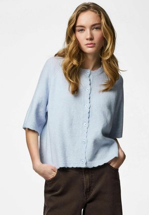 Fiatal nő hullámos szőke hajjal, világoskék gombos, rövid ujjú kardigánt és sötétbarna nadrágot visel, kezét a zsebében tartva.