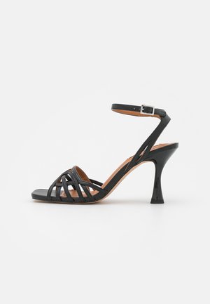 Czarna sandały na obcasie z paskowym wzorem, otworem na palce i regulowanym zapięciem na kostkę. Posiadają błyszczącą teksturę i konturowany obcas dla wsparcia.