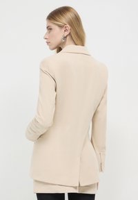 Blazer beige a coste con rever strutturato, spacco singolo sul retro e maniche lunghe. Realizzato in tessuto testurizzato, presenta una vestibilità sartoriale.