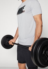 T-shirt Nike gris clair à manches courtes avec un grand logo noir, associé à un short noir. Individu tenant une barre de poids noire avec des plaques rondes.
