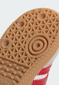 Rubber shoe sole in tan with a textured diamond pattern, circular accents, and ridges; features red and white stripe detailing on the side.