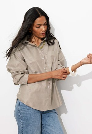 Femme aux longs cheveux foncés portant une chemise beige rayée à boutons et un jean bleu, ajustant un bracelet blanc à son poignet sur un fond blanc.