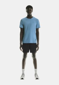 T-shirt de sport bleu clair, à manches courtes, col rond, associé à un short noir. Le mannequin porte des chaussettes blanches et des chaussures de sport grises.