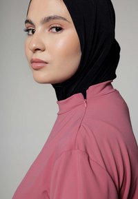 Roze hoge kraag kledingstuk met opgezette mouwen, voorzien van een rits aan de achterkant en gedragen met een zwarte hijab. Glad textuur en aansluitende silhoeet.