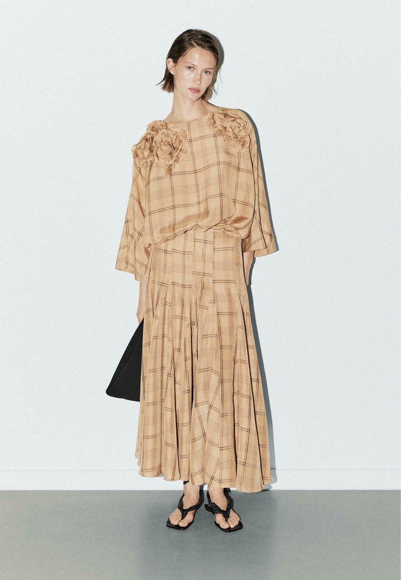 Beige geruite jurk met gerimpelde accenten op de schouders, vloeiende rok, korte mouwen en zwarte sandalen. De details creëren textuur en beweging.