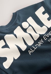 Primer plano de una sudadera azul oscuro con un gran texto blanco que dice "SMILE" y un texto más pequeño debajo que dice "ALL DAY EVERY".