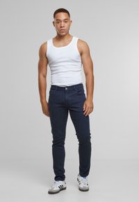 Tmavě modré skinny džíny s hladkou texturou a designem s pěti kapsami, spárované s bílým tílkem a bílými teniskami s černými akcenty.
