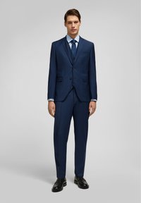 Costume ajusté bleu marine avec revers cranté, veste à un bouton, pantalon assorti, chemise habillée blanche et chaussures en cuir foncé.
