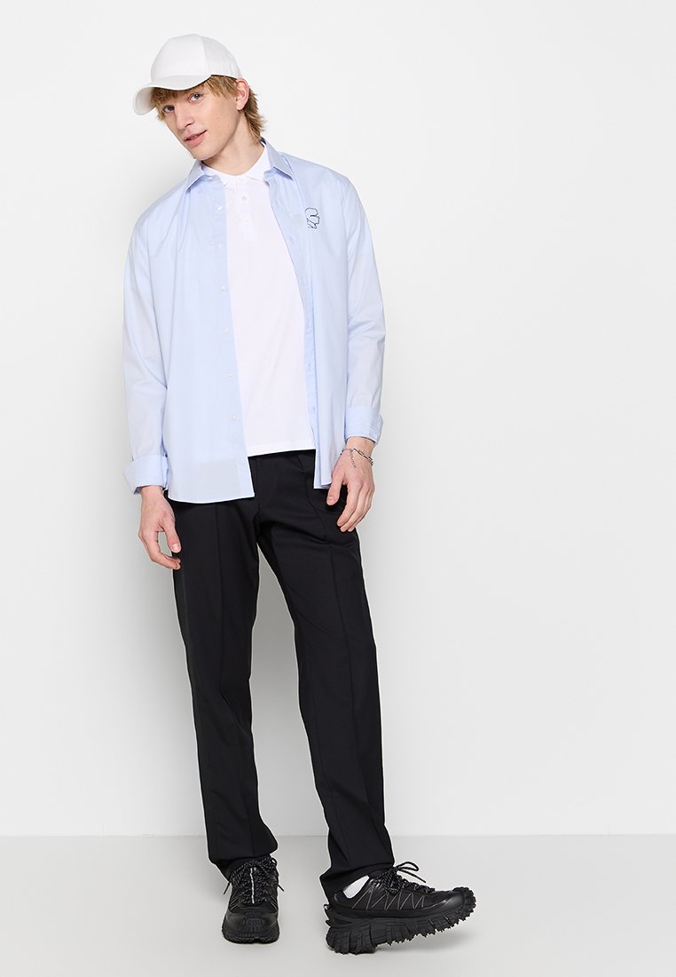 Himmelblaues Langarmhemd über einem weißen Polo, schwarze Hose und schwarze Sneakers mit weißen Akzenten. Eine weiße Kappe rundet das Outfit ab.