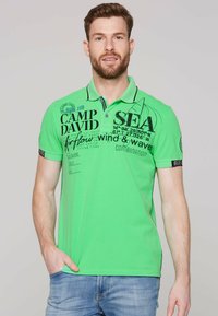 Zielona koszulka polo z krótkim rękawem, ozdobiona czarnym napisem graficznym oraz wzorem żaglówki. Posiada kołnierzyk oraz kontrastowe detale na rękawach.