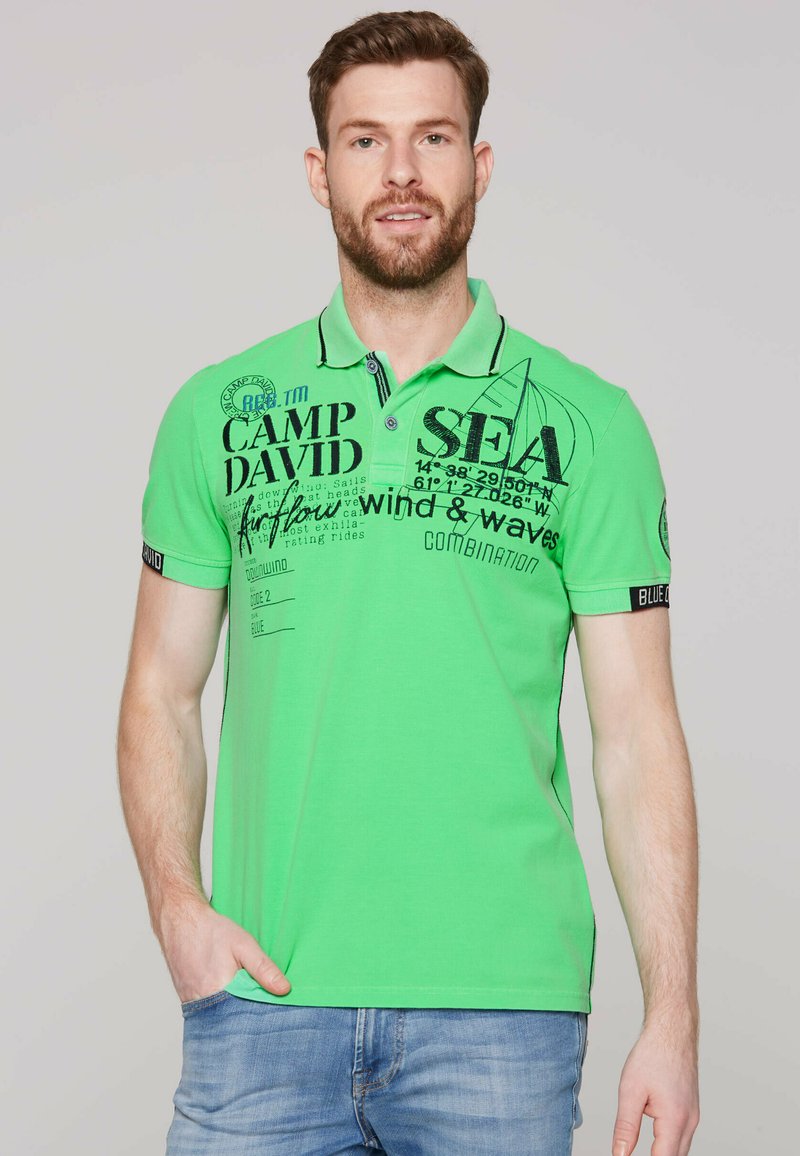 Zielona koszulka polo z krótkim rękawem, ozdobiona czarnym napisem graficznym oraz wzorem żaglówki. Posiada kołnierzyk oraz kontrastowe detale na rękawach.