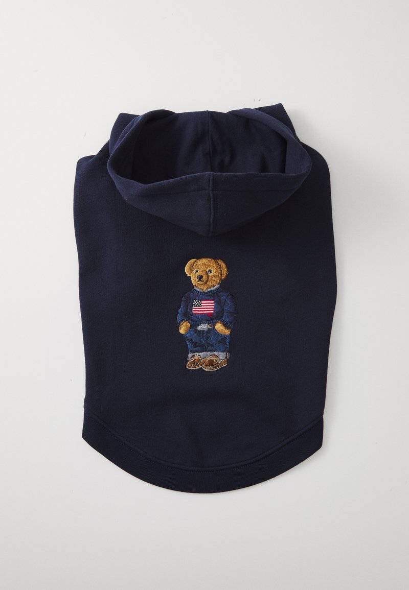 Sweat à capuche navy avec un ours en peluche brodé portant une chemise bleue avec un drapeau américain, un jean bleu et des chaussures brunes. Tissu doux, texture minimale.