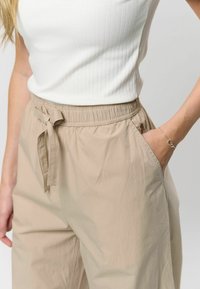 Béžové nohavice s elastickým pásom a detailom mašle, bočné vrecká a textúrovaná tkanina. Nosí sa s bielym rebrovaným tielkom.