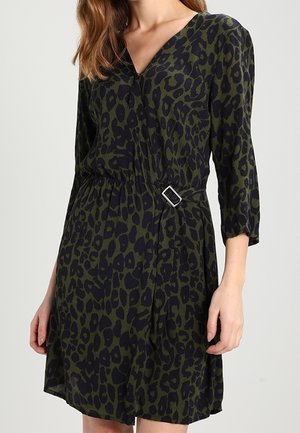 Frau trägt ein dunkelgrünes und schwarzes Leopardenmuster-Kleid mit Dreiviertelärmeln und einem seitlichen Schnallendetail an der Taille.
