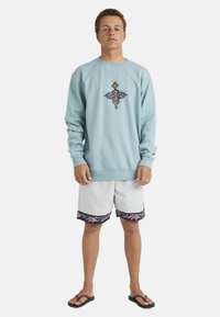 Young man standing wearing a light blue sweatshirt with orange-black emblem, white floral-hem shorts, and black flip-flops on a white background.